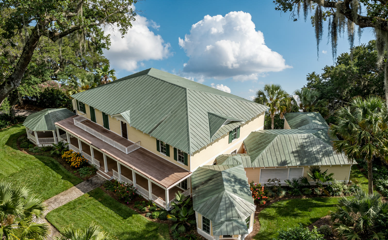 Aerial photograph of the house from above and to the
      south east. The green metal hipped roof fills the middle of the frame, the two storey porch runs along the
      near face, the gazebo sits at the far left and the diagonal wing runs off to the lower right, all in a
      garden of palms and live oaks.